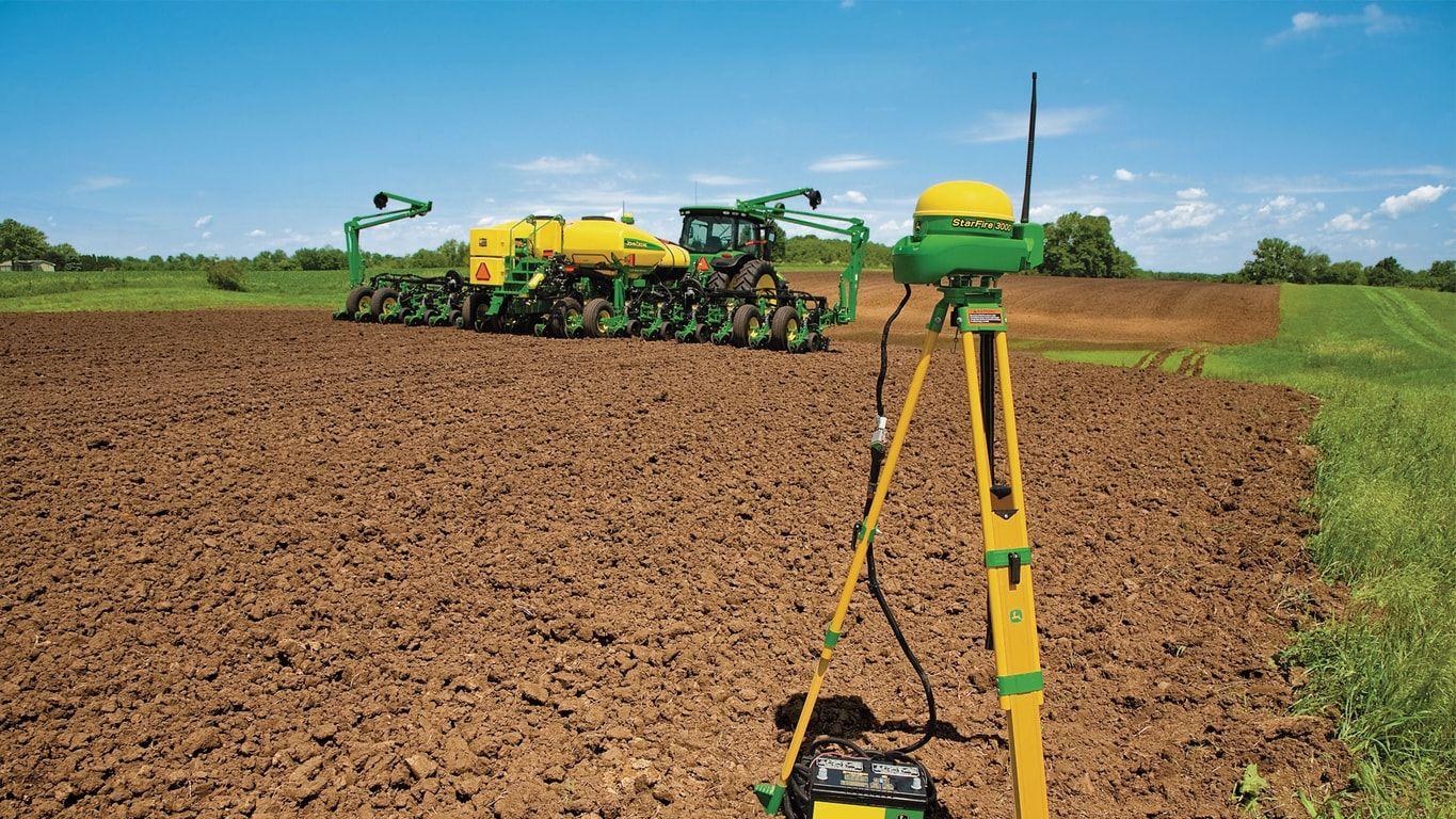 A GPS device in the foreground with a tractor planting crops in a large, freshly plowed field.