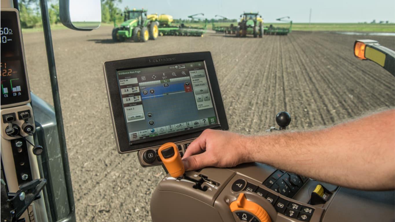 Farmer uses touchscreen in tractor cab showing field data and controls, with field work visible.