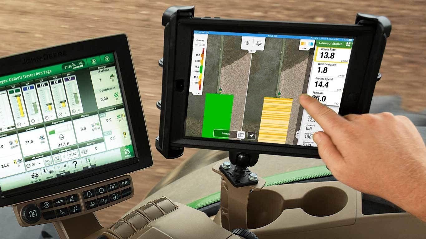 Person uses tablet and monitor to control farm machinery in a field with data on screens.