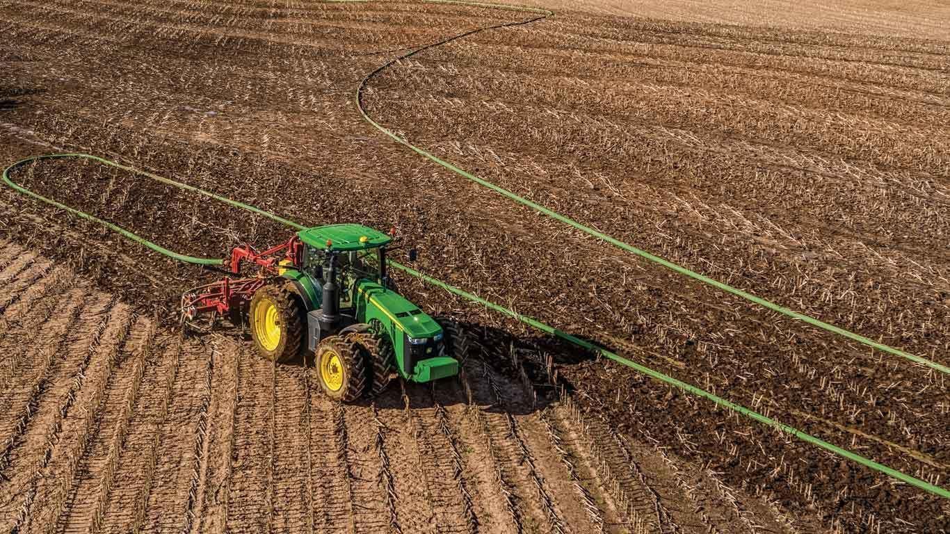 Green tractor with yellow wheels plows a large brown field with a long hose attached.